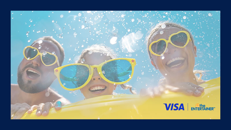 Three people smiling underwater, wearing oversized yellow sunglasses. Bubbles surround them in the bright blue water. Logos of Visa and The Entertainer are visible.
