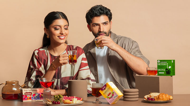 A man and woman enjoy tea in a warm-toned setting. They smile while holding teacups. Boxes of tea and a plate of croissants and fruit are on the table, conveying a cozy, inviting atmosphere.
