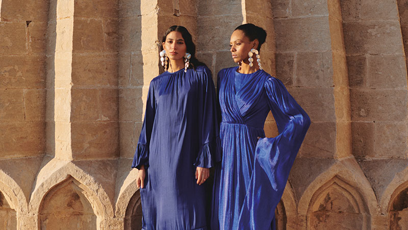 Two women in elegant blue dresses stand against a textured, sunlit stone wall. They wear large statement earrings, projecting a serene, stylish demeanor.