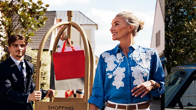 Woman in blue floral blouse smiles while standing outdoors beside a shopping bag-laden cart pushed by a suited man. The scene conveys joy and luxury.