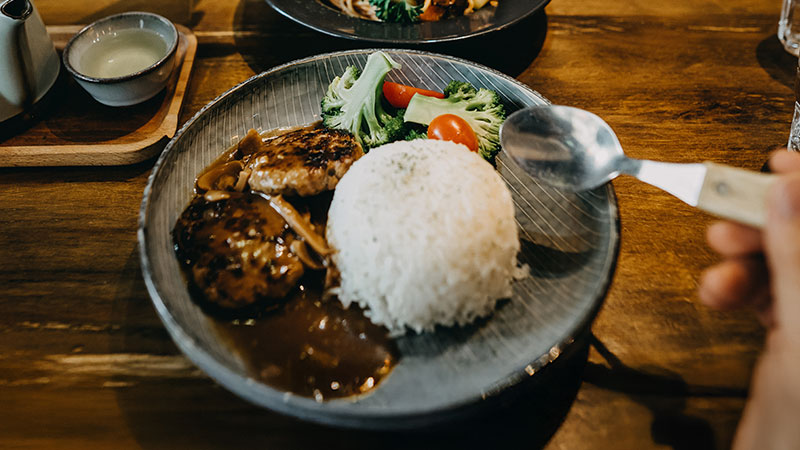 A hand holding a spoon approaches a plate with grilled hamburger patties, brown sauce, steamed rice, and a garnish of broccoli and cherry tomatoes.
