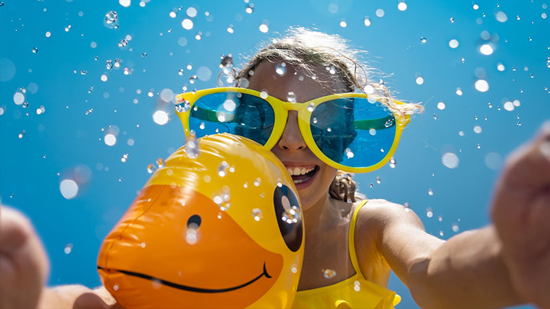 A child in oversized yellow sunglasses and a yellow swimsuit smiles joyfully while holding an inflatable duck in a pool, surrounded by splashing water under a clear blue sky.