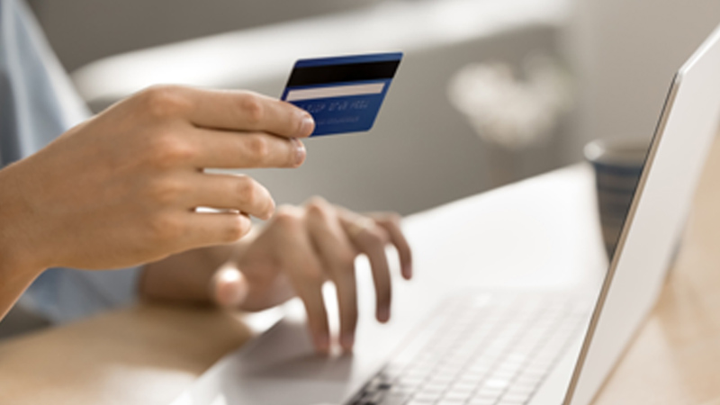 A person holds a blue credit card while typing on a laptop, suggesting online shopping or internet banking in a bright, cozy setting.