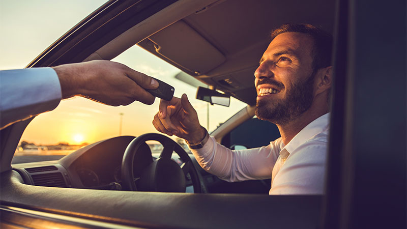 A man sitting in the driver’s seat of a car smiles as he receives a car key from another person through the open window at sunset.