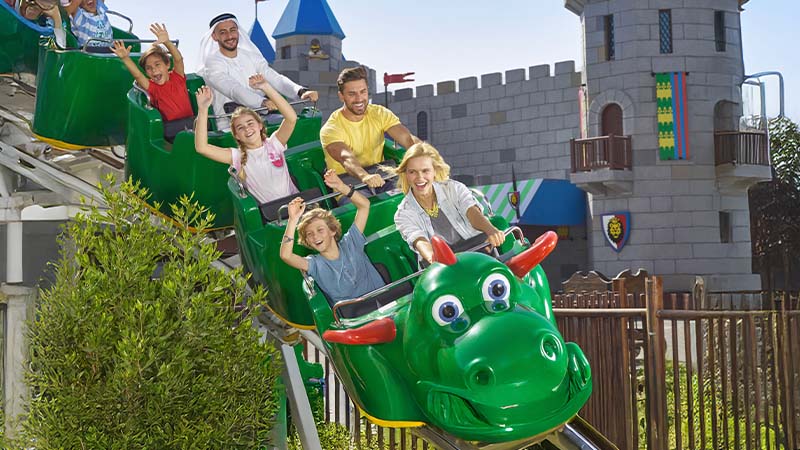 People riding a green dragon-themed roller coaster, smiling and raising their hands, with a castle structure in the background.