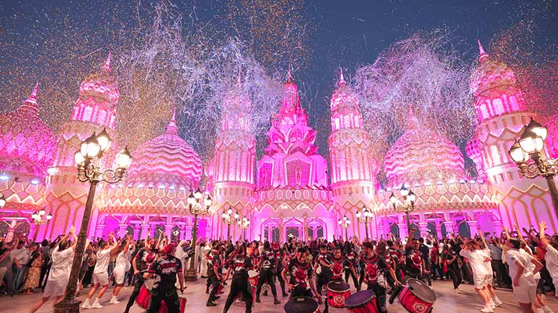 A large crowd celebrates in front of an ornate, pink-lit building with domes, with performers dancing and fireworks and confetti in the sky.
