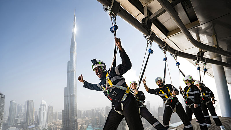 A group of people in safety harnesses participate in an outdoor skywalk activity on a tall building, with the Burj Khalifa visible in the background.