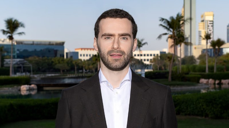 Man in a dark blazer and white shirt standing outdoors with palm trees and city buildings in the background