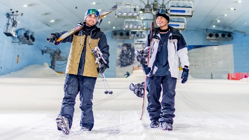 Two snowboarders walking indoors with their gear on a snowy slope