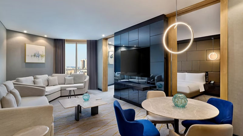 Modern hotel suite with a cozy living area featuring a white sectional sofa, circular dining table with blue chairs, and a sleek bedroom visible through a partition.