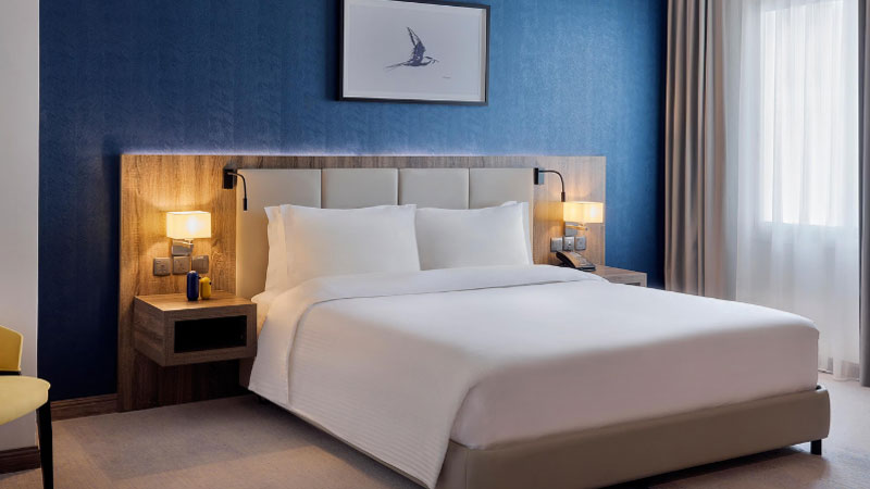 Cozy hotel room with a modern design. A large bed with white linens is flanked by wooden nightstands and lamps. A blue wall features artwork above.