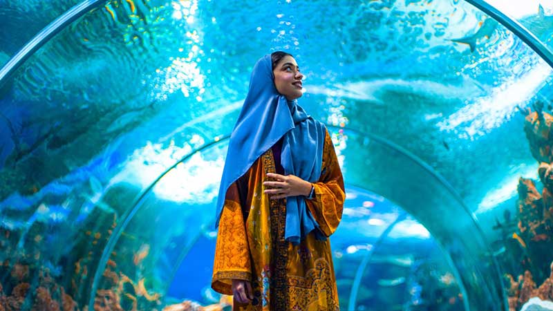 A woman in a colorful dress and blue hijab stands in an underwater tunnel, surrounded by vibrant marine life.